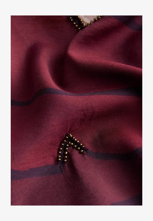 Közeli kép ráncos sötétvörös anyagról fekete vízszintes csíkokkal és kis rézből készült gyöngydíszítéssel, amely részleges háromszöget alkot.