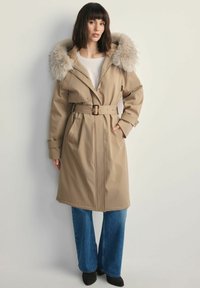 Beżowy trencz z paskiem, z futrzanym kapturem i szerokimi klapami, z bocznymi kieszeniami i gładką teksturą materiału. Połączony z dżinsami z szerokimi nogawkami.