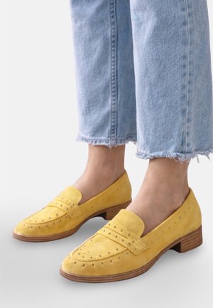 Pieds portant des mocassins en daim jaune avec des perforations décoratives et des petits talons en bois, associés à un jean bleu clair raccourci.