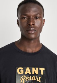Camiseta de algodón negra con un gran gráfico que tiene el texto "GANT Resort" en amarillo, cuello redondo y mangas cortas.