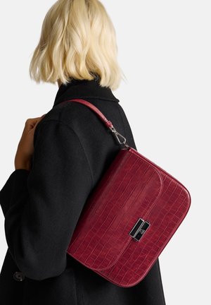 Borsa a spalla in pelle cremisi con una texture a motivo coccodrillo, forma rettangolare e chiusura in argento, indossata sopra un cappotto nero.