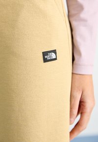 Beige textúrájú anyagból készült jogger nadrág, a combján fekete "The North Face" logóval, melyek testhez álló kialakítással és látható varrásokkal rendelkeznek.