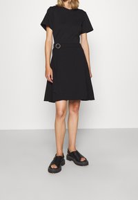 Czarna krótka sukienka z krótkim rękawem, z dopasowaną talią, z okrągłym paskiem jako akcentem. Gładki materiał i rozkloszowany dół, w zestawie z czarnymi butami na платформie.