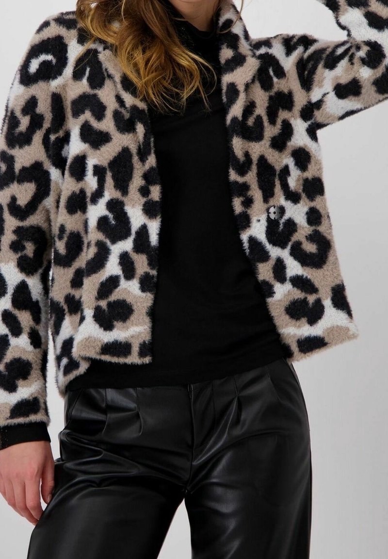 Femme portant une veste en peluche beige et noire à imprimé léopard par-dessus un haut noir, accompagnée d'un pantalon en similicuir noir.