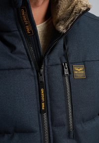 Námornícky modrá puffová bunda s golierom podšitým umelou kožušinou, dvojitým zipsom a značkou PME Legend. Obsahuje textúrovaný povrch.