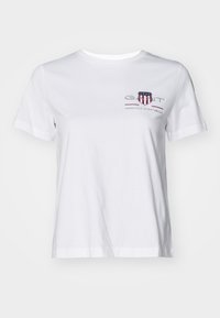 T-shirt bianco in cotone con maniche corte, colletto rotondo e un logo ricamato rosso e blu sul lato sinistro. Tessuto liscio, vestibilità morbida.