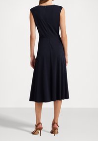 Marinblå ärmlös klänning med en figurnära överdel och utsvängd kjol. Har en dragkedja bak och en mjuk struktur. Bärs med bruna högklackade sandaler.