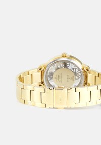 Gold-toned stainless steel watch with a round case, transparent back, and link bracelet. Features a smooth bezel and engraved logo clasp.