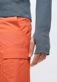 Camisola cinza de manga comprida com punhos alongados combinada com calças leves laranja, com bolso lateral e textura lisa.