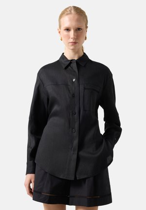 Kvinde med blondt hår iført en sort langærmet skjorte med knapper og sorte shorts med subtile udskæringsdetaljer, poserer med den ene hånd i lommen.