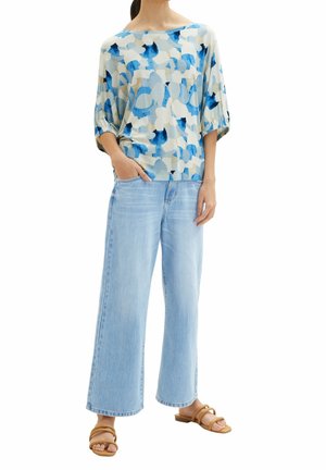 Femme portant un haut ample à motifs bleu et beige, un jean large bleu clair et des sandales marron à deux brides, debout avec une main dans la poche.