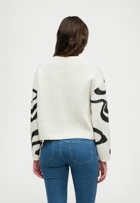 Nertas megztinis su tekstūruotu baltu kūnu ir juodais abstrakčiais raštais rankovėse. Yra trumpas dizainas ir raukšlėtas apatinis kraštas.