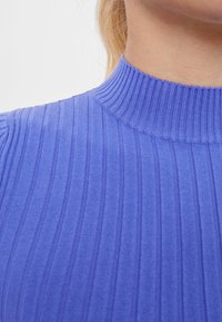 Top de punto acanalado en azul vibrante con cuello alto. El material tiene una textura suave y presenta rayas verticales a lo largo del cuerpo.