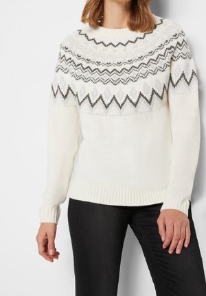 Femme portant un pull en maille blanc avec des motifs géométriques en zigzag gris sur la poitrine et le haut des manches, associée à un pantalon noir.