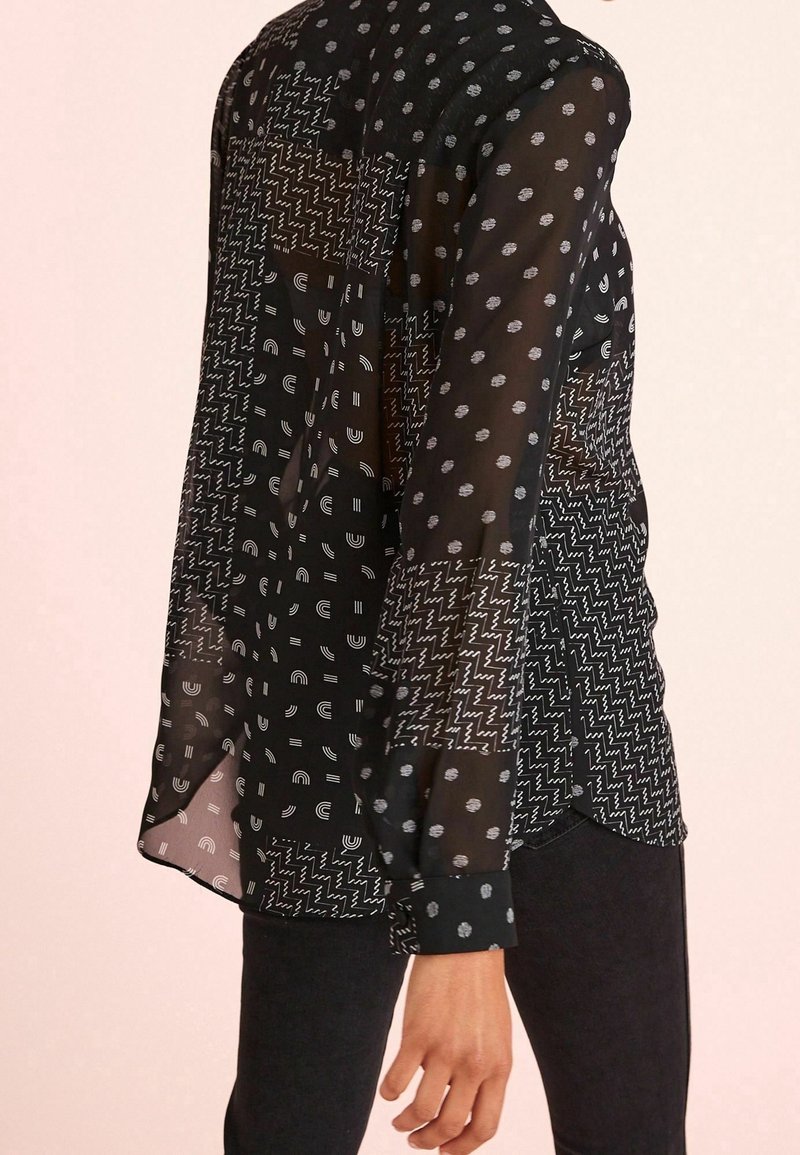 Blouse noire transparente avec motifs géométriques blancs mélangés, manches longues et poignets boutonnés, portée avec un pantalon noir.