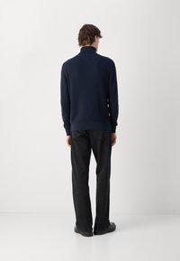 Homme debout, de dos, portant un pull texturé bleu marine, un jean noir et des chaussures noires, sur un fond clair uni.