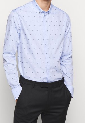 Homme portant une chemise à manches longues bleu clair avec de petits logos bleu foncé et un pantalon noir, mains dans les poches, sur fond blanc.