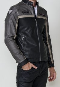 Homme portant une veste en cuir zippée noire et grise avec des écussons sur la poitrine et l'épaule, main dans la poche, sur un fond uni.