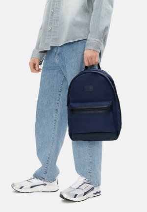 Osoba nosící světle modré džíny, pruhované tričko, bílé tenisky, držící tmavě modrý batoh za rukojeť na bílém pozadí.