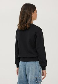 Felpa nera in cotone con scollo a girocollo, polsini a costine e vestibilità comoda, abbinata a pantaloni cargo in denim blu. Vista posteriore mostrata.