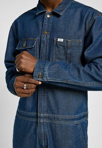 Homme fermant le poignet d'une veste en denim bleu foncé Lee avec des boutons en métal et des poches poitrine, portant deux bagues en argent à la main gauche.
