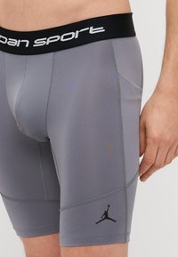 Sive kratke sportske hlače s crnim pojasom na kojem je ispisan tekst "Jordan Sport." Uključuju crni logo i detalj bočne džepne.