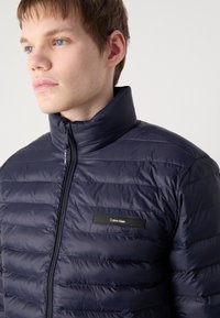Navy puffer jacket with a high collar, horizontal quilted pattern, and a black Calvin Klein logo patch on the chest. Smooth, shiny texture.