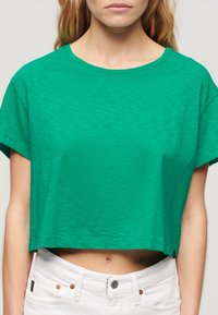Grøn cropped t-shirt lavet af letvægts, tekstureret stof. Har en rund halsudskæring og korte ærmer, parret med hvidt taljebånd.