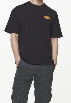 Schwarzes Baumwoll-T-Shirt mit kurzen Ärmeln, ausgestattet mit einem kleinen roten und gelben Logo auf der Brust. Kombiniert mit grauen Cargo-Hosen.