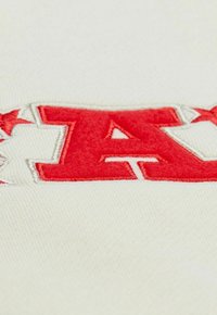Rote und weiße bestickte Buchstabe "A" mit Sternen auf weißem, strukturiertem Stoff, zeigt detaillierte Stickerei und Gewebe.