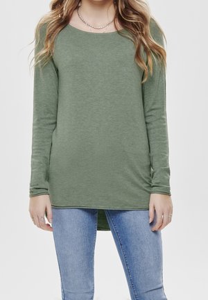 Maglia verde a maniche lunghe realizzata in tessuto morbido, con scollatura rotonda e orlo posteriore leggermente più lungo, abbinata a jeans blu chiaro.