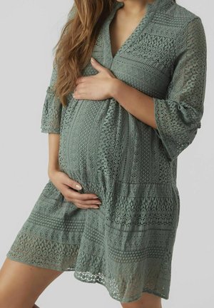 Schwangere Frau in grünem Spitzenkleid, die sanft mit beiden Händen ihren Bauch vor einem einfachen Hintergrund hält.