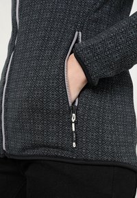 Hand binnenin het ritssluitingvak van een zwarte jas met een grijs geometrisch patroon en een witte ritsschlinger aan de zijkant.