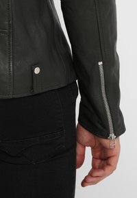 Veste en cuir vert foncé au design ajusté, avec des poignets zippés et un détail de bouton-pression à l'ourlet. Fini texturé et doux.