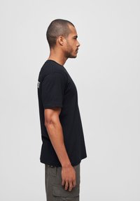 Črna kratka majica s kratkimi rokavi in krožnim izrezom, prikazana na osebi s strani. Ima ohlapen kroj in mehko bombažno teksturo.