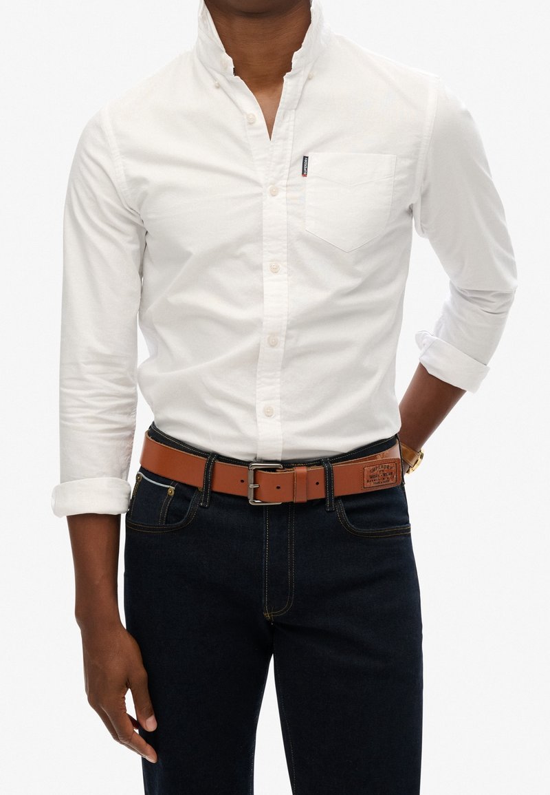 Personne portant une chemise blanche à manches longues avec les poignets retroussés, un jean bleu foncé et une ceinture en cuir marron avec une boucle rectangulaire.