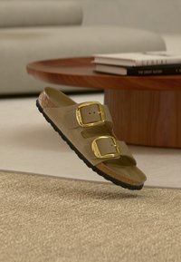 Μπεζ σανδάλι με δύο μεγάλες χρυσές αγκράφες, πάτος από φελλό, μαύρη σόλα, να πλανάται πάνω από μπεζ χαλί σε μοντέρνο σαλόνι.