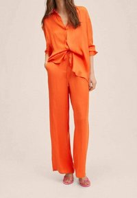 Světle oranžový dvoudílný outfit složený z volné košile s knoflíky a kalhot s šňůrkou. Spárováno s růžovými sandály. Hladká, texturovaná tkanina.