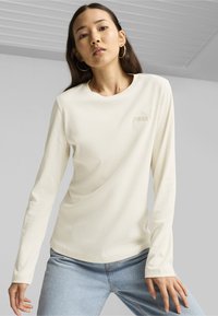 Dlouhý rukáv, ribovaný top v off-white barvě s kulatým výstřihem a jemným logem. V kombinaci se světle modrými džíny, na neutrálním pozadí.