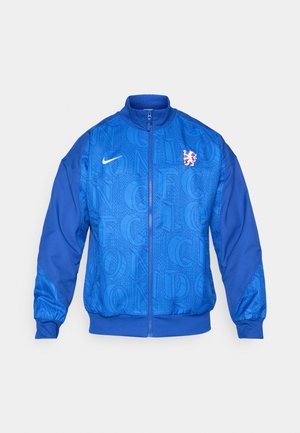 Modrá sportovní bunda na zip s logem Nike, erb lva Chelsea FC a opakovaným vzorem textu "LONDON" na přední straně a rukávech.