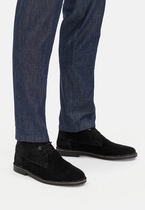 Schwarze Chukka-Stiefel aus Wildleder mit Schnürdesign, ausgestattet mit tonalen Nähten und einer flachen Gummisohle, kombiniert mit dunklen Jeanshosen.