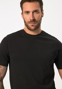 Černé tričko s kulatým výstřihem vyrobené z hladké, lehké látky. Krátké rukávy a přiléhavý střih. Viditelný detail tetování na levém rameni.