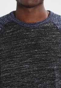 Pull en maille gris foncé avec un corps texturé, des manches raglan bleu contrastant et un col rond. Tissu doux et décontracté, idéal pour le superposition.