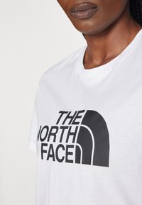 Bijela pamučna majica s velikim crnim logotipom koji glasi "THE NORTH FACE." Klasični izrez oko vrata i kratki rukavi.