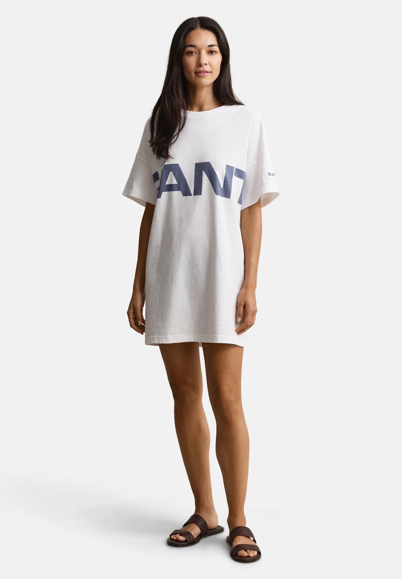Femme debout portant une robe T-shirt blanche oversize avec « GANT » en grandes lettres bleues et des sandales plates tressées marron foncé.