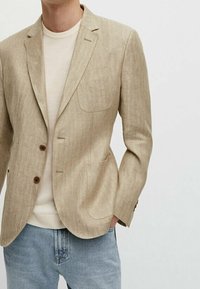 Beżowa lniana marynarka z klapą z wycięciem, trzema guzikami i dwoma przednimi kieszeniami, noszona na kremowej koszuli, w połączeniu z jasnoniebieskimi dżinsami.