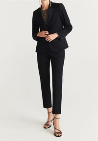 Completo nero su misura con una vestibilità slim, caratterizzato da una giacca con revers e pantaloni affusolati. Indossato con un top testurizzato e tacchi con cinturini.