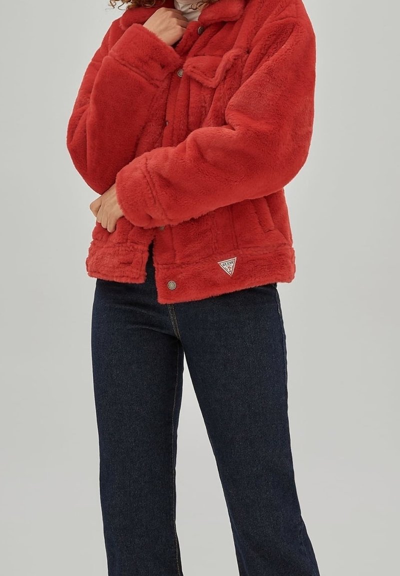 Rode faux fur jas met een kraag en knoopsluiting, voorzien van twee voorzakken en een logo-patch aan de zoom. Gecombineerd met donkere jeans.