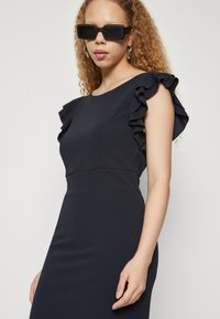 Temno modra obleka s prirezano krojenjem in ruffles rokavi, izdelana iz gladkega blaga. Model nosi črne pravokotne sončna očala in viseče srebrne uhane.