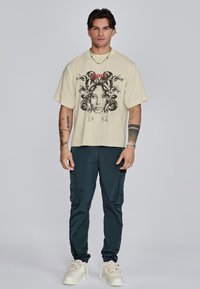 Crème kortemouwen T-shirt met een grafische print van een gezicht en slangen; gecombineerd met donkere cargobroeken en lichtgekleurde sneakers.